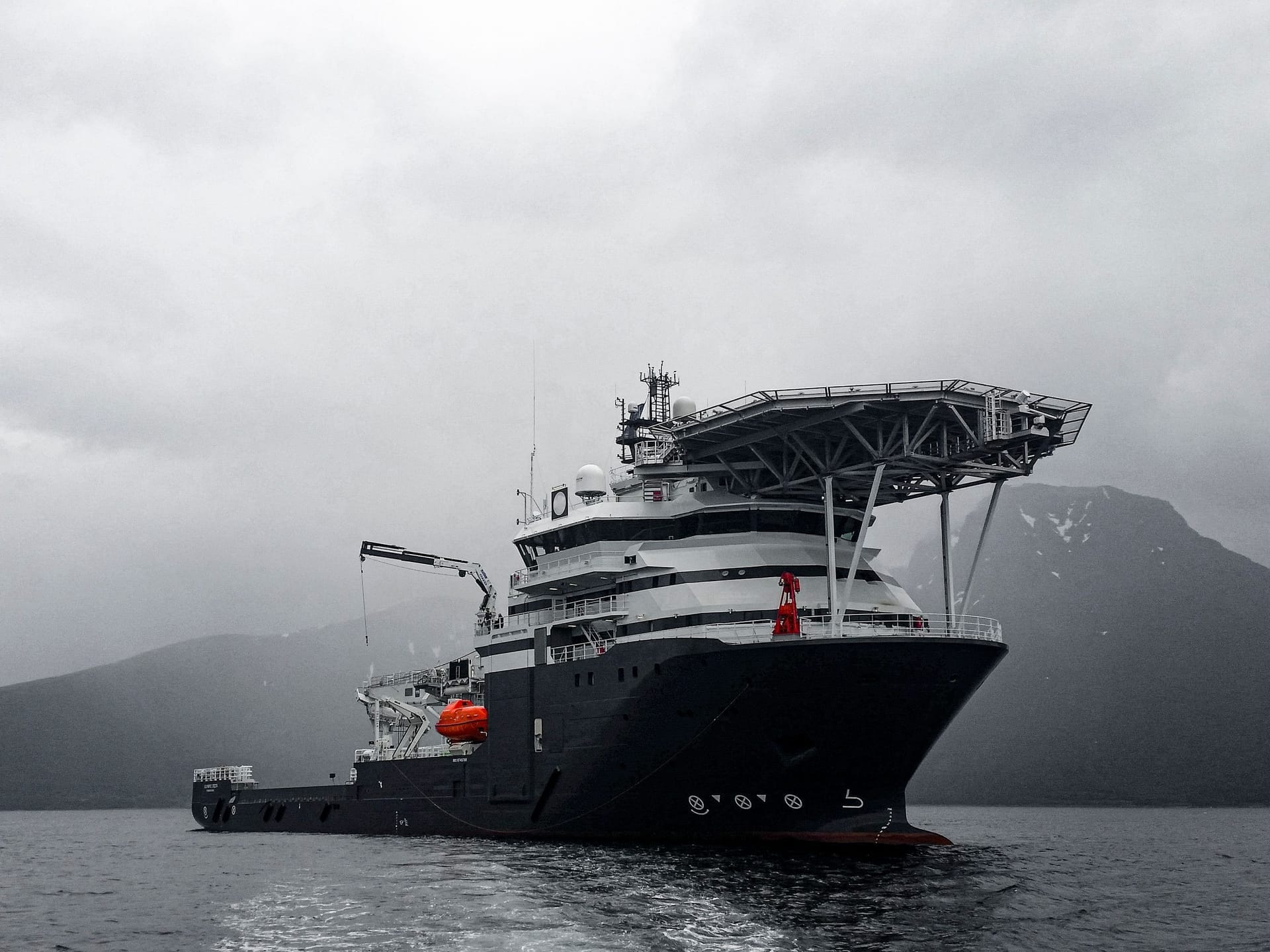 Mørkt offshorefartøy med helikopterdekk til ankers i ein disig fjord